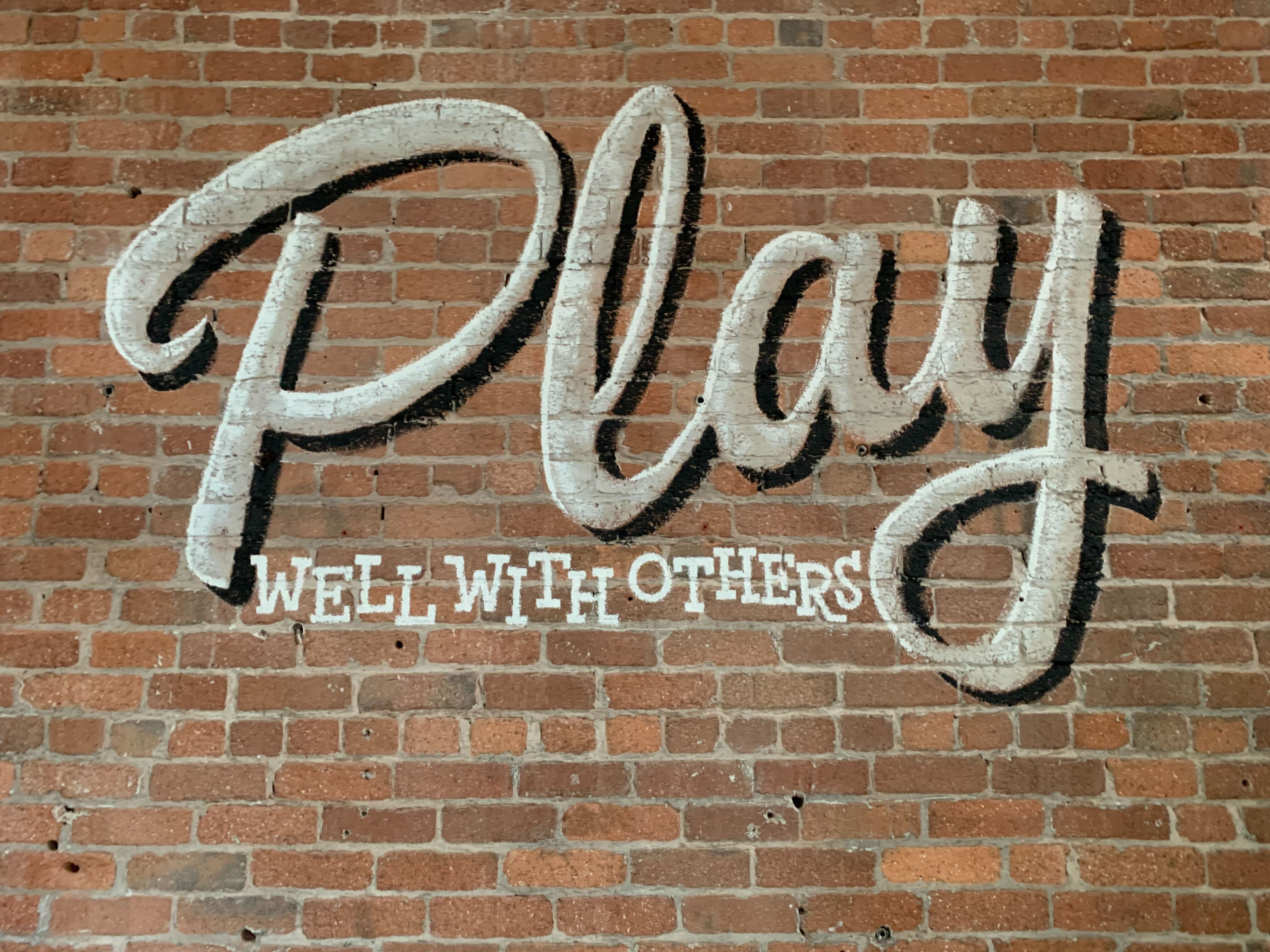 A sign on a brick wall that reads "Play well with others"